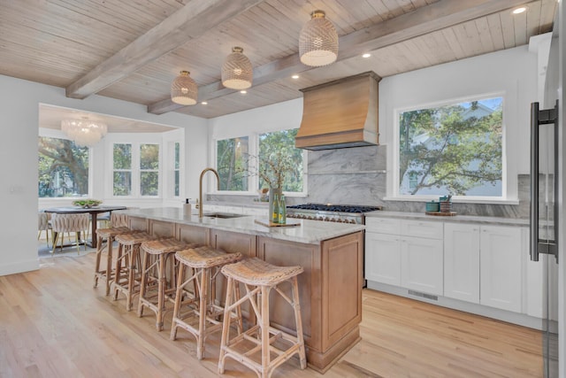 kitchen with custom range hood, white cabinets, sink, a kitchen island with sink, and light stone counters