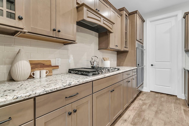 kitchen with premium range hood, tasteful backsplash, stainless steel gas cooktop, light stone countertops, and light wood-type flooring