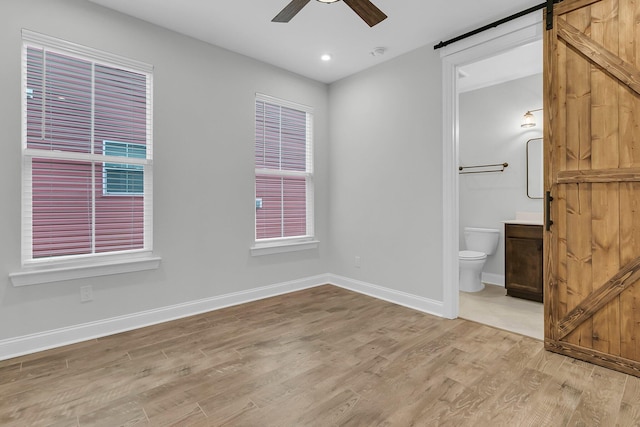 unfurnished bedroom with ceiling fan, ensuite bath, a barn door, and light wood-type flooring