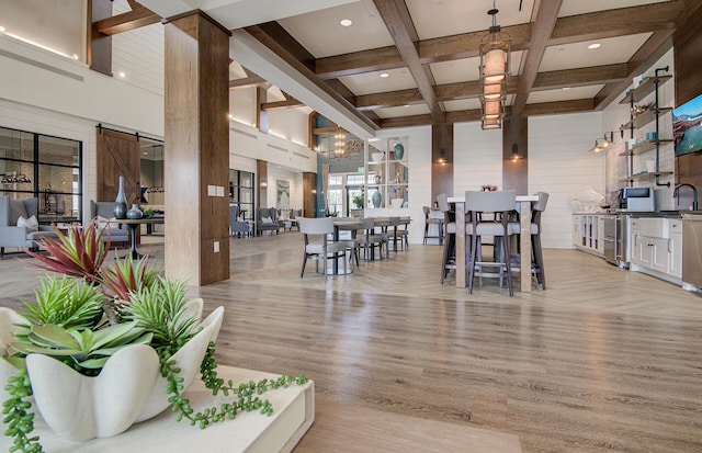 dining space featuring light hardwood / wood-style flooring, beam ceiling, a high ceiling, coffered ceiling, and a barn door