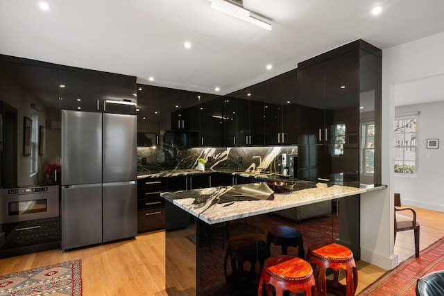 kitchen with light stone counters, stainless steel appliances, light wood-type flooring, backsplash, and a kitchen breakfast bar