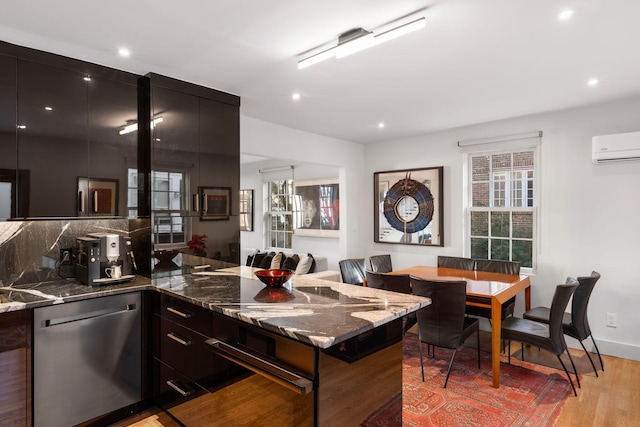 kitchen with a wall mounted AC, stainless steel dishwasher, light hardwood / wood-style floors, and dark stone countertops