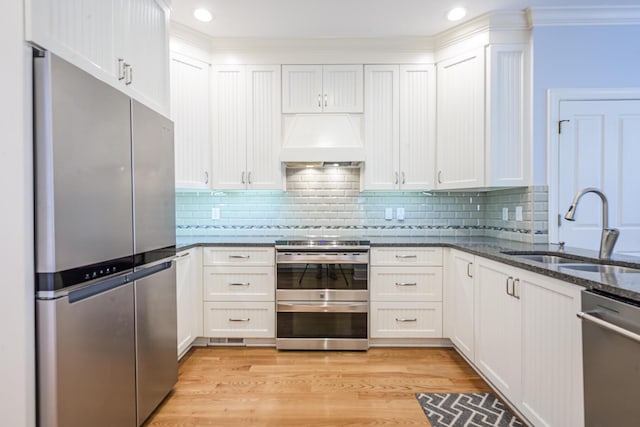 kitchen with stainless steel appliances, premium range hood, dark stone countertops, a sink, and light wood-type flooring