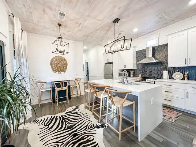 kitchen with pendant lighting, wall chimney exhaust hood, sink, stainless steel refrigerator, and a center island with sink