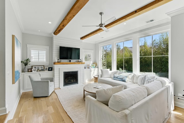 living area featuring ceiling fan, a large fireplace, beam ceiling, ornamental molding, and light wood-style flooring