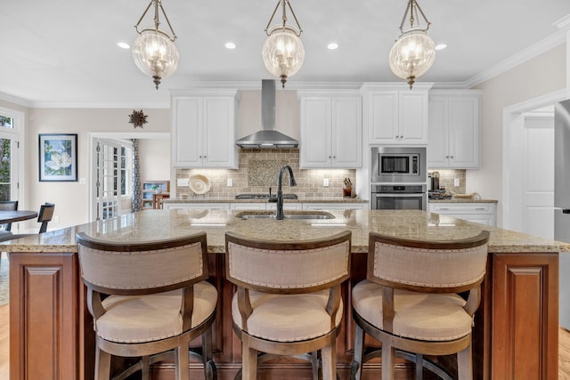 kitchen featuring wall chimney exhaust hood, appliances with stainless steel finishes, ornamental molding, a kitchen island with sink, and a sink