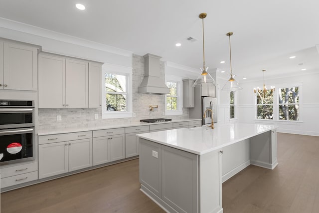 kitchen featuring appliances with stainless steel finishes, decorative light fixtures, decorative backsplash, a kitchen island with sink, and custom range hood