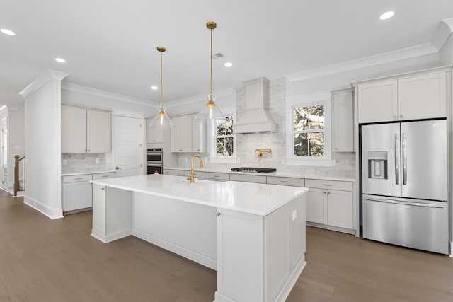 kitchen featuring crown molding, appliances with stainless steel finishes, white cabinetry, custom range hood, and an island with sink