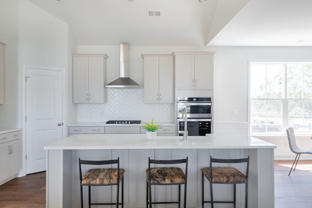 kitchen featuring dark wood-type flooring, stainless steel double oven, wall chimney range hood, and an island with sink