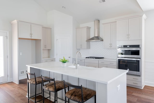kitchen featuring wall chimney range hood, double oven, a kitchen island with sink, and light wood-type flooring