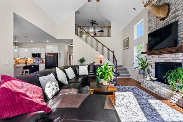 living room with stairway, a ceiling fan, a brick fireplace, high vaulted ceiling, and wood finished floors