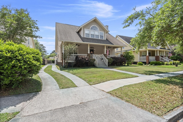 bungalow-style home featuring covered porch, ceiling fan, a chimney, roof with shingles, and a front lawn