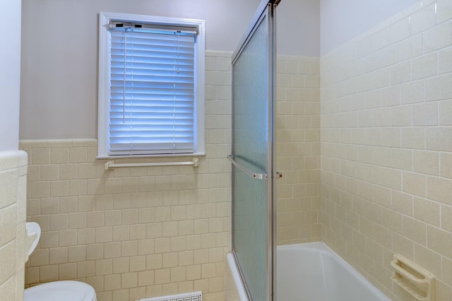 bathroom with combined bath / shower with glass door, wainscoting, toilet, and tile walls