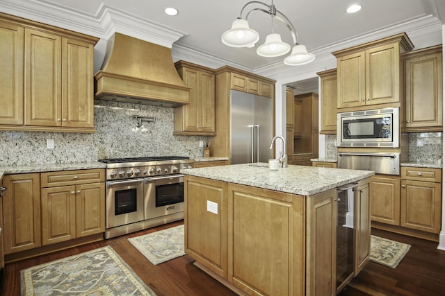 kitchen featuring wall chimney exhaust hood, light stone counters, built in appliances, an island with sink, and pendant lighting