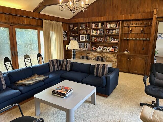 living room featuring built in features, wood walls, lofted ceiling with beams, a chandelier, and light colored carpet