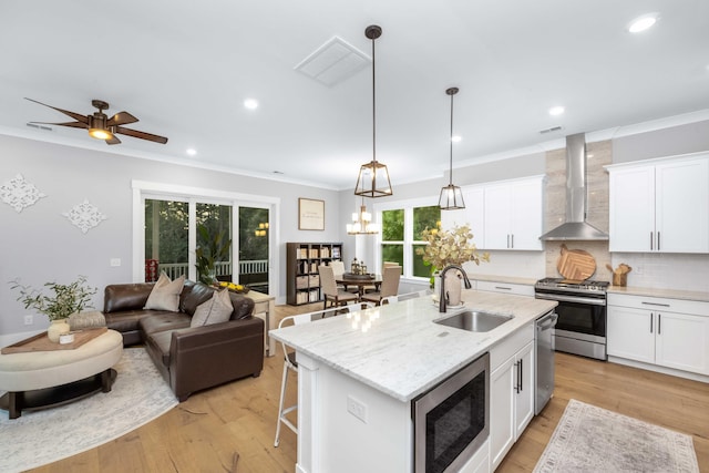 kitchen featuring appliances with stainless steel finishes, hanging light fixtures, a kitchen island with sink, white cabinets, and wall chimney range hood