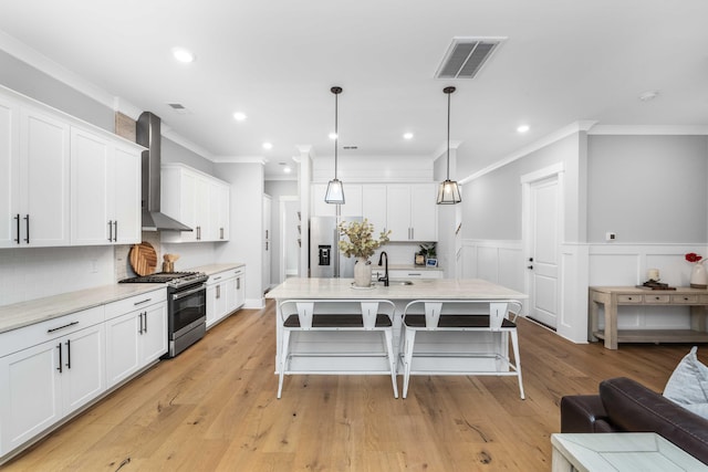 kitchen with light hardwood / wood-style floors, appliances with stainless steel finishes, wall chimney exhaust hood, an island with sink, and hanging light fixtures