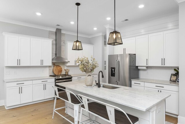 kitchen with a center island with sink, white cabinetry, wall chimney range hood, appliances with stainless steel finishes, and decorative light fixtures