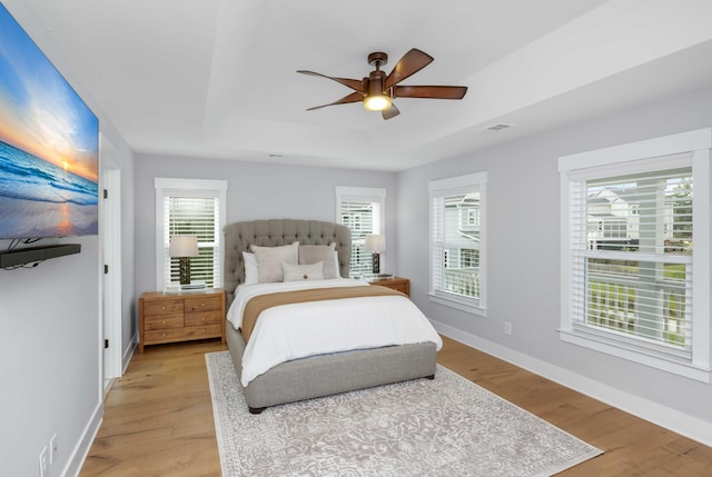 bedroom with ceiling fan, multiple windows, a raised ceiling, and light hardwood / wood-style flooring