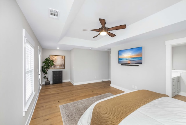 bedroom with ensuite bathroom, light hardwood / wood-style flooring, ceiling fan, and a raised ceiling