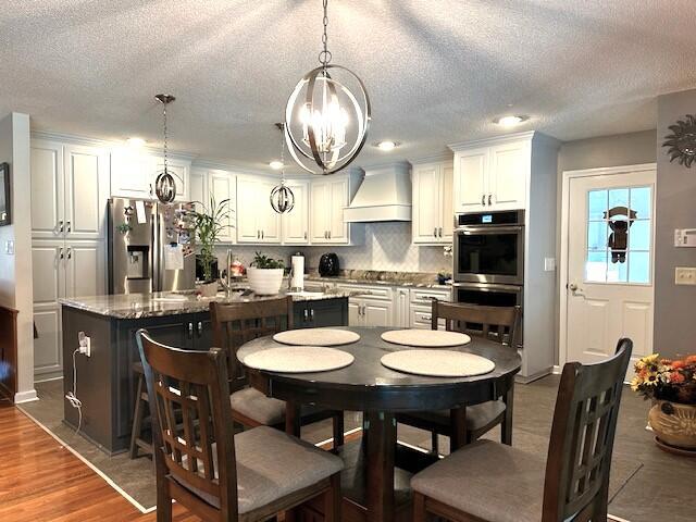 kitchen with wood finished floors, custom exhaust hood, an island with sink, stainless steel appliances, and a notable chandelier