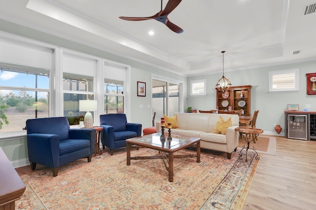 living room with wine cooler, light hardwood / wood-style flooring, a tray ceiling, ceiling fan with notable chandelier, and ornamental molding