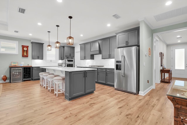 kitchen with gray cabinetry, custom range hood, stainless steel appliances, pendant lighting, and a center island with sink