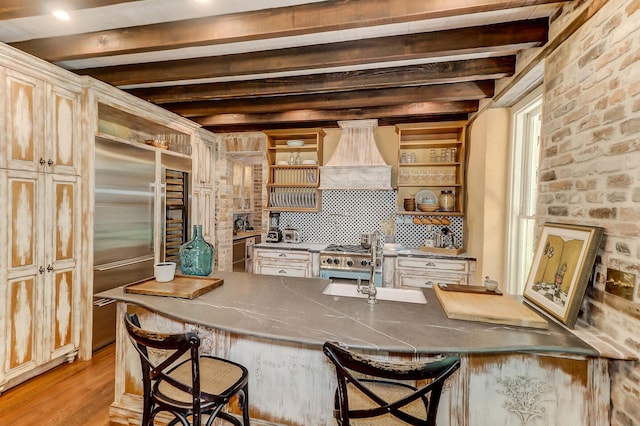kitchen with beamed ceiling, stainless steel built in refrigerator, a breakfast bar area, custom exhaust hood, and light hardwood / wood-style floors