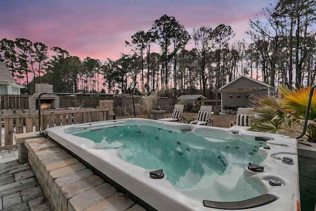 pool at dusk featuring an outdoor structure, fence, and a hot tub