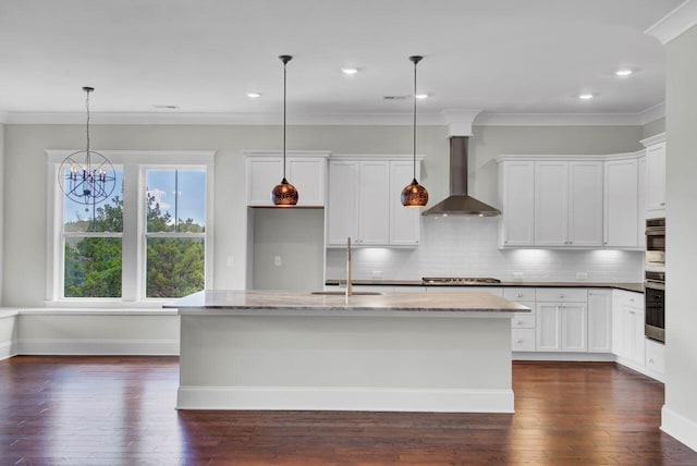 kitchen with a center island with sink, white cabinets, hanging light fixtures, wall chimney exhaust hood, and light stone counters