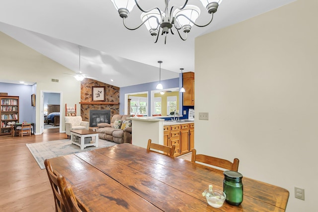 dining area featuring light wood-style flooring, ceiling fan with notable chandelier, visible vents, a stone fireplace, and vaulted ceiling