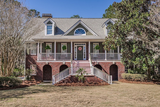 coastal inspired home with stairway, covered porch, a front yard, a shingled roof, and brick siding