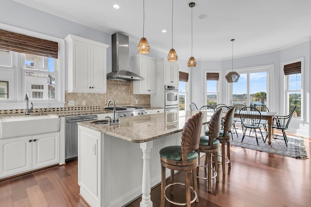 kitchen featuring wall chimney range hood, sink, an island with sink, appliances with stainless steel finishes, and white cabinetry