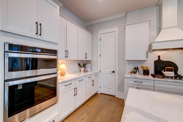 kitchen with custom exhaust hood, light hardwood / wood-style flooring, double oven, white cabinets, and backsplash