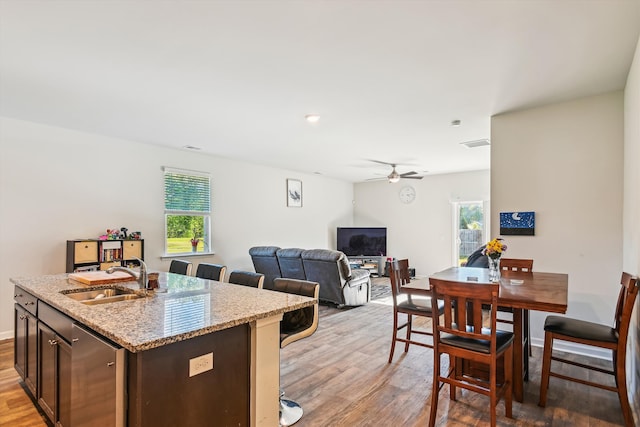 kitchen with a sink, a center island with sink, light wood-style floors, a ceiling fan, and light stone countertops