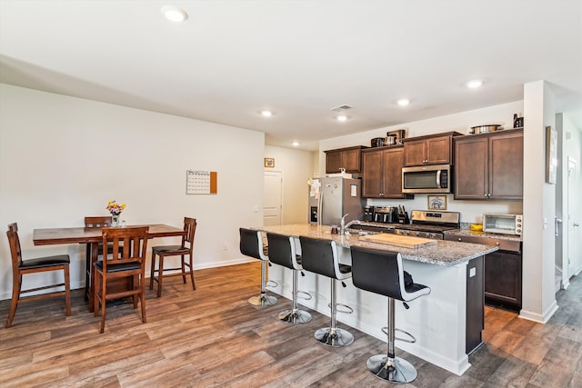 kitchen featuring an island with sink, stainless steel appliances, a breakfast bar, dark brown cabinets, and wood finished floors