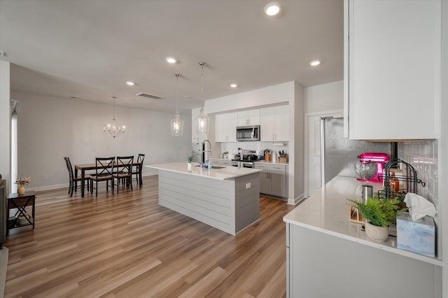 kitchen with hanging light fixtures, appliances with stainless steel finishes, white cabinetry, a center island with sink, and light hardwood / wood-style floors