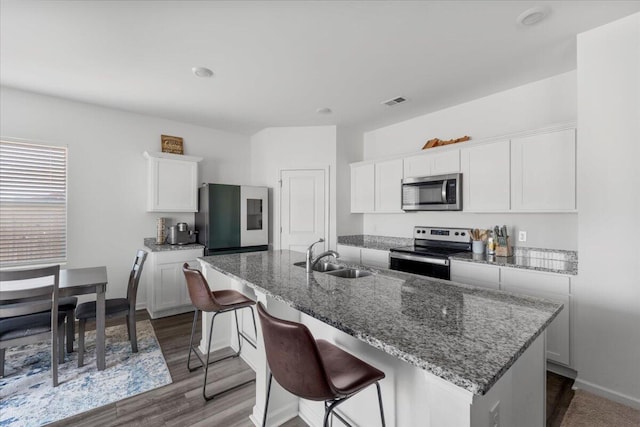 kitchen with light stone countertops, a kitchen island with sink, a sink, appliances with stainless steel finishes, and white cabinetry