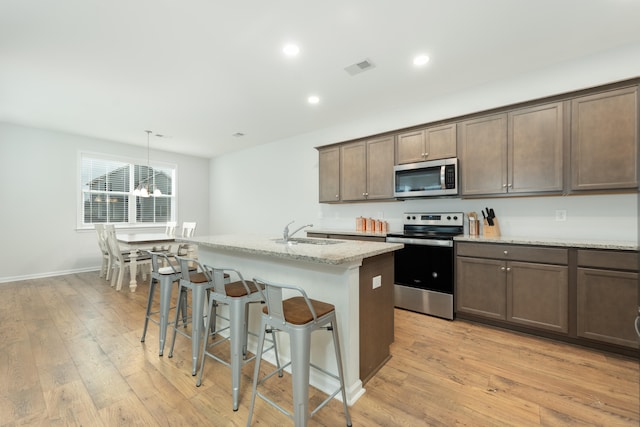 kitchen with appliances with stainless steel finishes, light wood-type flooring, decorative light fixtures, a kitchen island with sink, and sink