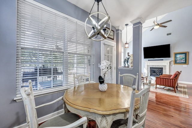 dining space with ceiling fan with notable chandelier, a fireplace, wood finished floors, visible vents, and decorative columns