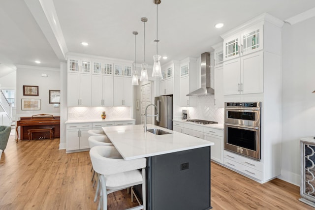 kitchen featuring wall chimney range hood, sink, white cabinetry, a kitchen island with sink, and stainless steel appliances