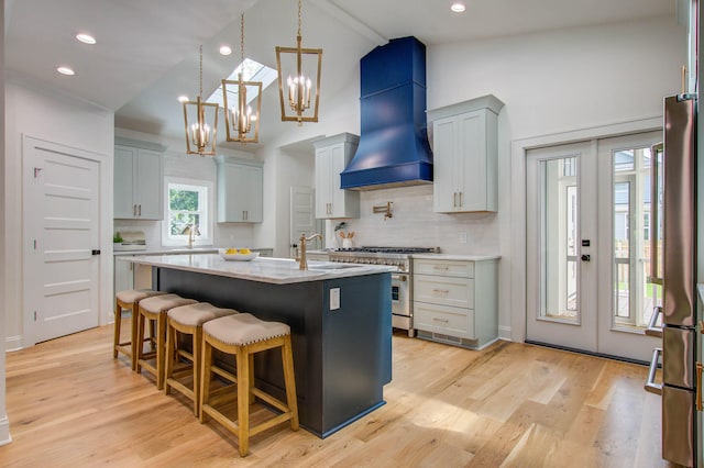 kitchen featuring custom exhaust hood, high vaulted ceiling, a center island with sink, light wood-type flooring, and appliances with stainless steel finishes