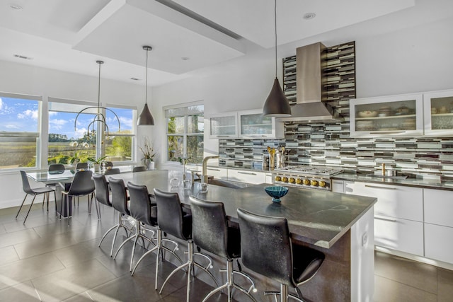 kitchen with a center island with sink, decorative backsplash, white cabinetry, wall chimney range hood, and stove