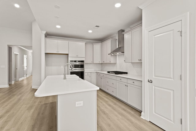 kitchen with decorative backsplash, wall chimney exhaust hood, light wood-style floors, stainless steel appliances, and a kitchen island with sink