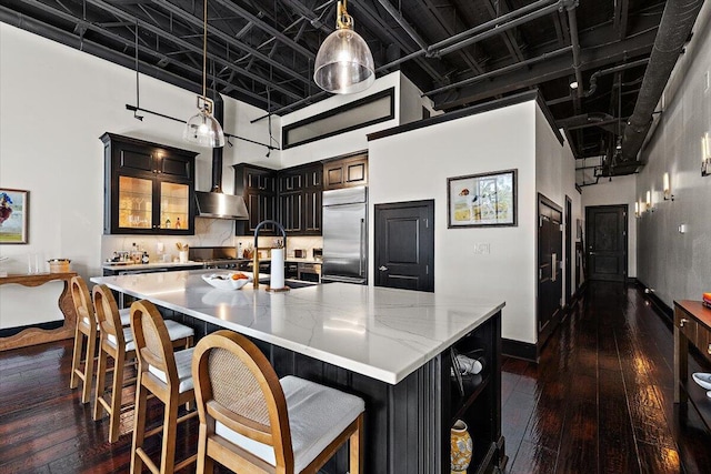 kitchen with dark wood-style flooring, a towering ceiling, a large island with sink, built in fridge, and wall chimney exhaust hood