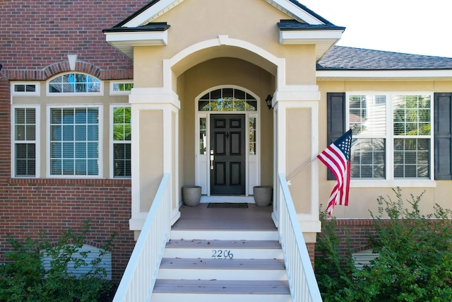 entrance to property with stucco siding, brick siding, and a shingled roof