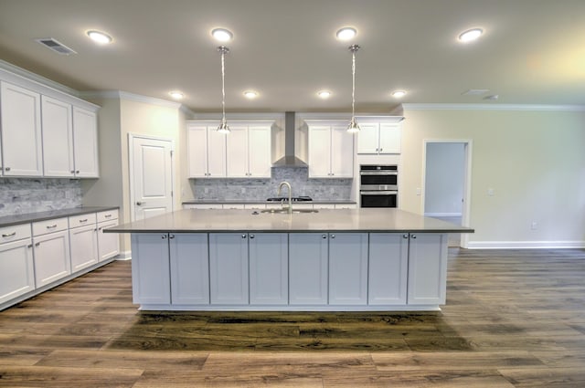 kitchen with pendant lighting, wall chimney range hood, white cabinetry, tasteful backsplash, and a center island with sink