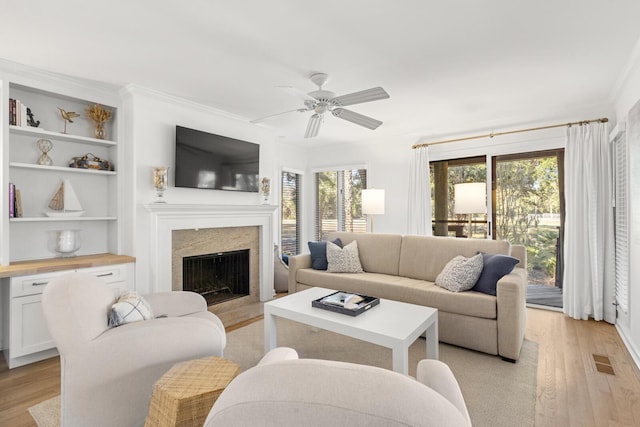 living room with built in features, a high end fireplace, ceiling fan, crown molding, and light wood-type flooring