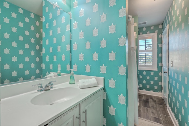 bathroom with wood finish floors, visible vents, vanity, baseboards, and wallpapered walls