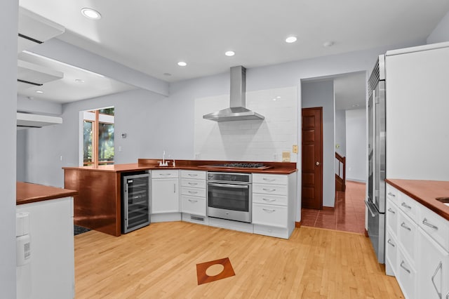 kitchen featuring wall chimney range hood, decorative backsplash, light wood-type flooring, appliances with stainless steel finishes, and beverage cooler